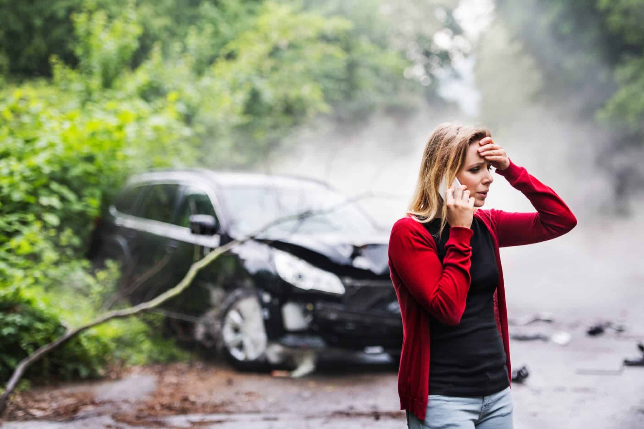 une femme est au téléphone avec son assurance auto. Sa voiture est endommagée par un arbre